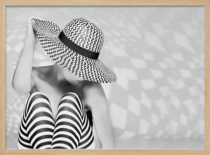A black and white artistic photograph of a woman sitting down, her face obscured by a large, wide-brimmed sun hat with a black and white chevron pattern. She is wearing black and white striped leggings and holding the brim of her hat with one hand. The image is set against a plain background with patterned shadows and is presented within a silver frame. Artwork