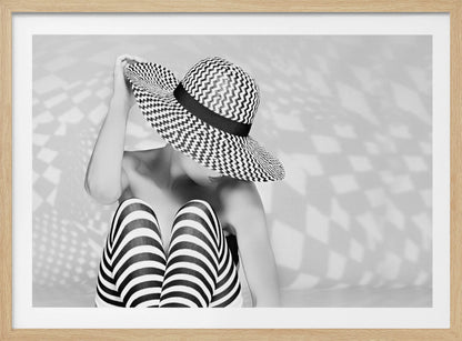 A black and white artistic photograph of a woman sitting down, her face obscured by a large, wide-brimmed sun hat with a black and white chevron pattern. She is wearing black and white striped leggings and holding the brim of her hat with one hand. The image is set against a plain background with patterned shadows and is presented within a silver frame. Artwork