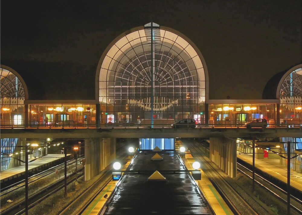 A symmetrical view of a modern train station at night, with a large illuminated arched glass window. Below, empty, wet train tracks and platforms reflect the station's lights, while cars cross an overpass in the middle ground. Wall Art