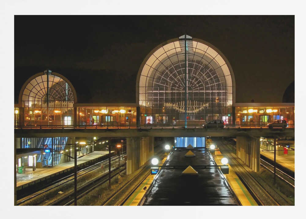 A symmetrical view of a modern train station at night, with a large illuminated arched glass window. Below, empty, wet train tracks and platforms reflect the station's lights, while cars cross an overpass in the middle ground. Wall Art