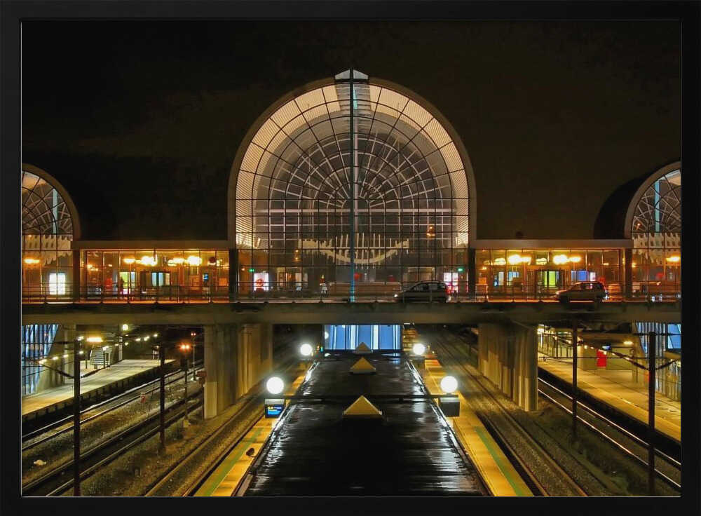 A symmetrical view of a modern train station at night, with a large illuminated arched glass window. Below, empty, wet train tracks and platforms reflect the station's lights, while cars cross an overpass in the middle ground. Wall Art