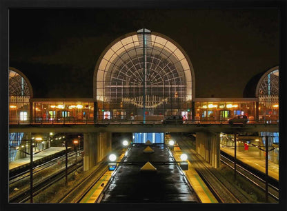 A symmetrical view of a modern train station at night, with a large illuminated arched glass window. Below, empty, wet train tracks and platforms reflect the station's lights, while cars cross an overpass in the middle ground. Wall Art