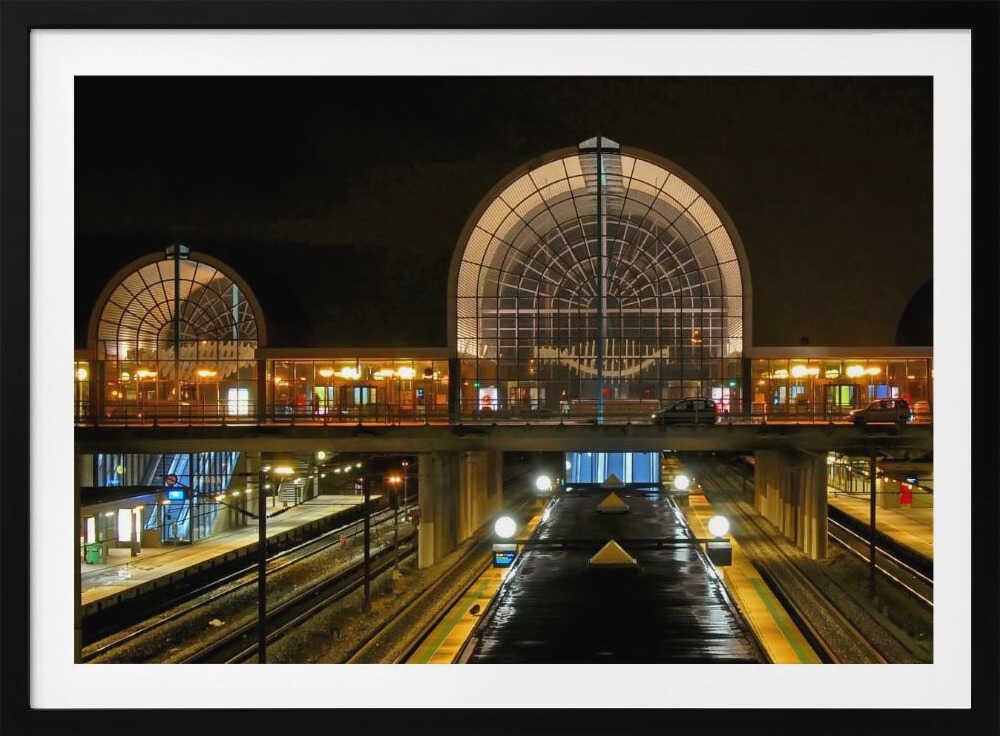 A symmetrical view of a modern train station at night, with a large illuminated arched glass window. Below, empty, wet train tracks and platforms reflect the station's lights, while cars cross an overpass in the middle ground. Wall Art