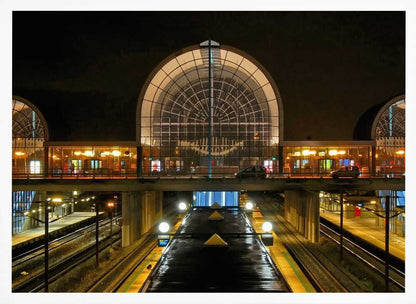 A symmetrical view of a modern train station at night, with a large illuminated arched glass window. Below, empty, wet train tracks and platforms reflect the station's lights, while cars cross an overpass in the middle ground. Wall Art