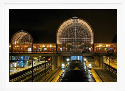 A symmetrical view of a modern train station at night, with a large illuminated arched glass window. Below, empty, wet train tracks and platforms reflect the station's lights, while cars cross an overpass in the middle ground. Wall Art
