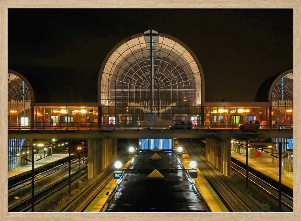 A symmetrical view of a modern train station at night, with a large illuminated arched glass window. Below, empty, wet train tracks and platforms reflect the station's lights, while cars cross an overpass in the middle ground. Wall Art