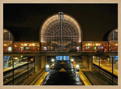 A symmetrical view of a modern train station at night, with a large illuminated arched glass window. Below, empty, wet train tracks and platforms reflect the station's lights, while cars cross an overpass in the middle ground. Wall Art
