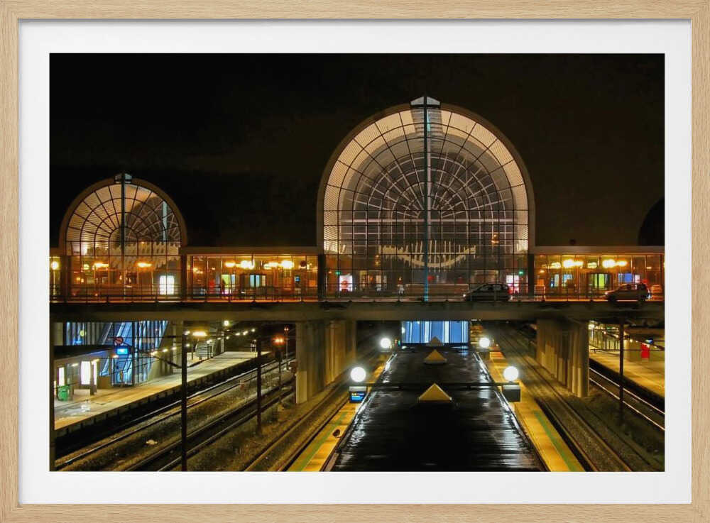 A symmetrical view of a modern train station at night, with a large illuminated arched glass window. Below, empty, wet train tracks and platforms reflect the station's lights, while cars cross an overpass in the middle ground. Wall Art