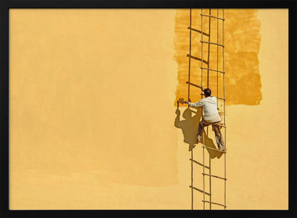 A man viewed from behind stands on a rope ladder, painting a large, bright yellow wall a slightly darker shade of yellow, casting a stark shadow in the bright sun. The image is presented within a silver frame. Decor