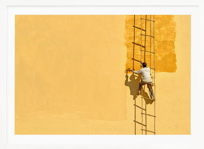 A man viewed from behind stands on a rope ladder, painting a large, bright yellow wall a slightly darker shade of yellow, casting a stark shadow in the bright sun. The image is presented within a silver frame. Decor