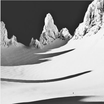 A framed, high-contrast black and white photograph of a majestic, snow-covered mountain range. Jagged peaks are sunlit against a stark black sky, casting long, dramatic shadows across a vast, pristine snowfield below. A tiny, solitary figure stands in the snow, highlighting the immense scale of the landscape. Print