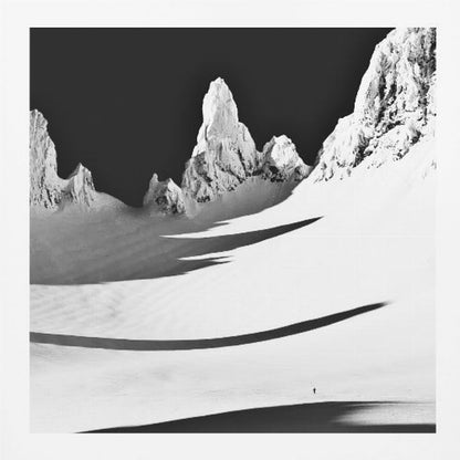 A framed, high-contrast black and white photograph of a majestic, snow-covered mountain range. Jagged peaks are sunlit against a stark black sky, casting long, dramatic shadows across a vast, pristine snowfield below. A tiny, solitary figure stands in the snow, highlighting the immense scale of the landscape. Print