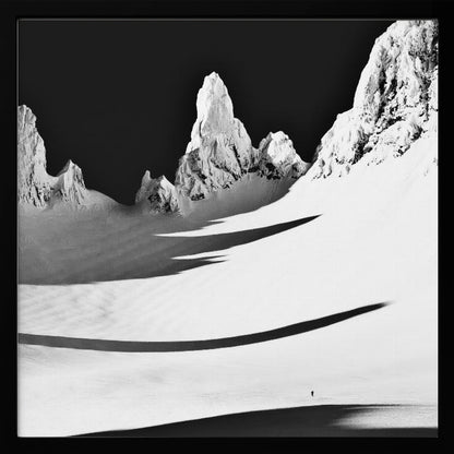 A framed, high-contrast black and white photograph of a majestic, snow-covered mountain range. Jagged peaks are sunlit against a stark black sky, casting long, dramatic shadows across a vast, pristine snowfield below. A tiny, solitary figure stands in the snow, highlighting the immense scale of the landscape. Print