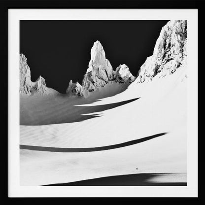 A framed, high-contrast black and white photograph of a majestic, snow-covered mountain range. Jagged peaks are sunlit against a stark black sky, casting long, dramatic shadows across a vast, pristine snowfield below. A tiny, solitary figure stands in the snow, highlighting the immense scale of the landscape. Print