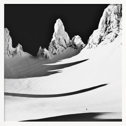 A framed, high-contrast black and white photograph of a majestic, snow-covered mountain range. Jagged peaks are sunlit against a stark black sky, casting long, dramatic shadows across a vast, pristine snowfield below. A tiny, solitary figure stands in the snow, highlighting the immense scale of the landscape. Print