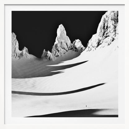 A framed, high-contrast black and white photograph of a majestic, snow-covered mountain range. Jagged peaks are sunlit against a stark black sky, casting long, dramatic shadows across a vast, pristine snowfield below. A tiny, solitary figure stands in the snow, highlighting the immense scale of the landscape. Print