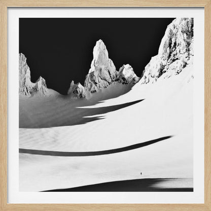 A framed, high-contrast black and white photograph of a majestic, snow-covered mountain range. Jagged peaks are sunlit against a stark black sky, casting long, dramatic shadows across a vast, pristine snowfield below. A tiny, solitary figure stands in the snow, highlighting the immense scale of the landscape. Print