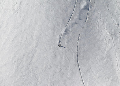 An aerial, top-down photograph of a lone snowboarder carving a turn down a vast, textured, snowy mountain slope, leaving fresh tracks in the powder. The image is presented in a simple, grey frame. Wall Art