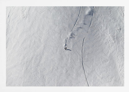 An aerial, top-down photograph of a lone snowboarder carving a turn down a vast, textured, snowy mountain slope, leaving fresh tracks in the powder. The image is presented in a simple, grey frame. Wall Art