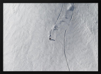 An aerial, top-down photograph of a lone snowboarder carving a turn down a vast, textured, snowy mountain slope, leaving fresh tracks in the powder. The image is presented in a simple, grey frame. Wall Art