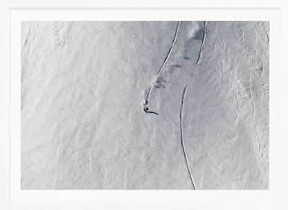 An aerial, top-down photograph of a lone snowboarder carving a turn down a vast, textured, snowy mountain slope, leaving fresh tracks in the powder. The image is presented in a simple, grey frame. Wall Art