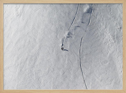 An aerial, top-down photograph of a lone snowboarder carving a turn down a vast, textured, snowy mountain slope, leaving fresh tracks in the powder. The image is presented in a simple, grey frame. Wall Art