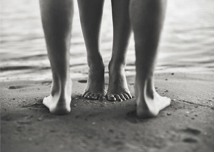 A framed, black and white photograph showing a low-angle view of two people's bare feet and lower legs standing on a wet sandy beach. The central pair of feet is in focus, while another person's legs in the foreground are blurred, creating depth. The water's edge is visible in the background. Artwork