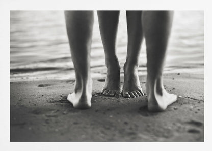 A framed, black and white photograph showing a low-angle view of two people's bare feet and lower legs standing on a wet sandy beach. The central pair of feet is in focus, while another person's legs in the foreground are blurred, creating depth. The water's edge is visible in the background. Artwork