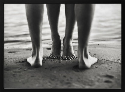 A framed, black and white photograph showing a low-angle view of two people's bare feet and lower legs standing on a wet sandy beach. The central pair of feet is in focus, while another person's legs in the foreground are blurred, creating depth. The water's edge is visible in the background. Artwork