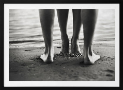 A framed, black and white photograph showing a low-angle view of two people's bare feet and lower legs standing on a wet sandy beach. The central pair of feet is in focus, while another person's legs in the foreground are blurred, creating depth. The water's edge is visible in the background. Artwork