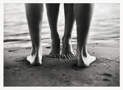 A framed, black and white photograph showing a low-angle view of two people's bare feet and lower legs standing on a wet sandy beach. The central pair of feet is in focus, while another person's legs in the foreground are blurred, creating depth. The water's edge is visible in the background. Artwork