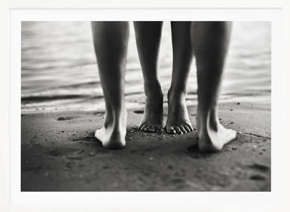 A framed, black and white photograph showing a low-angle view of two people's bare feet and lower legs standing on a wet sandy beach. The central pair of feet is in focus, while another person's legs in the foreground are blurred, creating depth. The water's edge is visible in the background. Artwork