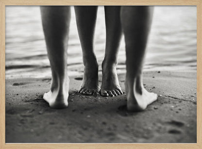 A framed, black and white photograph showing a low-angle view of two people's bare feet and lower legs standing on a wet sandy beach. The central pair of feet is in focus, while another person's legs in the foreground are blurred, creating depth. The water's edge is visible in the background. Artwork