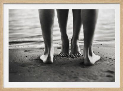 A framed, black and white photograph showing a low-angle view of two people's bare feet and lower legs standing on a wet sandy beach. The central pair of feet is in focus, while another person's legs in the foreground are blurred, creating depth. The water's edge is visible in the background. Artwork