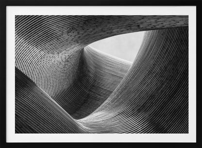 A framed, black and white abstract photograph of a swirling, textured structure. The low-angle shot looks up at curving walls made of thin, parallel lines, creating a dynamic sense of movement and depth, with an opening to a bright sky at the top. Artwork