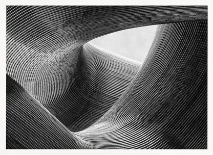 A framed, black and white abstract photograph of a swirling, textured structure. The low-angle shot looks up at curving walls made of thin, parallel lines, creating a dynamic sense of movement and depth, with an opening to a bright sky at the top. Artwork