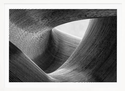 A framed, black and white abstract photograph of a swirling, textured structure. The low-angle shot looks up at curving walls made of thin, parallel lines, creating a dynamic sense of movement and depth, with an opening to a bright sky at the top. Artwork