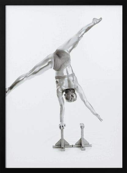 A black and white studio photograph of a muscular gymnast coated in silver paint, performing a dynamic one-armed handstand on a small block. Their legs are split wide apart in a display of strength and flexibility against a clean white background, all enclosed in a black frame. Decor