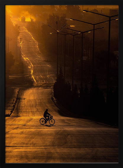 A silhouette of a person riding a bicycle across a wide, winding road illuminated by the warm, golden light of a sunrise or sunset. The road stretches uphill into the distance, and tall streetlights stand in shadow on the right. Artwork