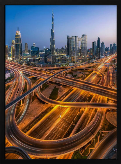 A long-exposure, aerial photograph of the Dubai skyline at dusk. The iconic Burj Khalifa stands tall in the center, surrounded by other illuminated skyscrapers against a deep blue twilight sky. In the foreground, a complex web of highways and overpasses glows with the orange and white light trails of moving traffic. Decor