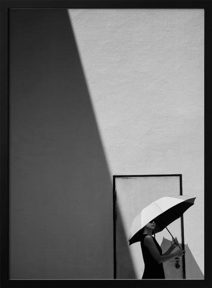 A minimalist black and white photograph of a woman in a black dress holding a two-toned umbrella. She stands in front of a door against a light wall, with a sharp, diagonal shadow dramatically cutting across the upper left of the image, creating a high-contrast, geometric composition. Decor