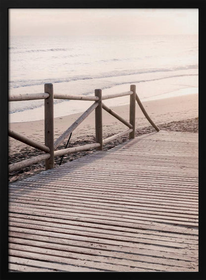 A wooden boardwalk with a rustic log fence leads down to a sandy beach with gentle waves lapping at the shore under a soft, hazy sunrise or sunset. Decor