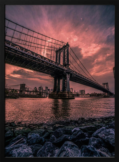 A dramatic low-angle view of the Manhattan Bridge over the East River at sunset. The sky is filled with pink and red clouds, which are reflected in the water below. The dark silhouette of the bridge contrasts with the vibrant sky, and the New York City skyline is visible in the background. In the foreground, dark, wet rocks line the riverbank. Decor