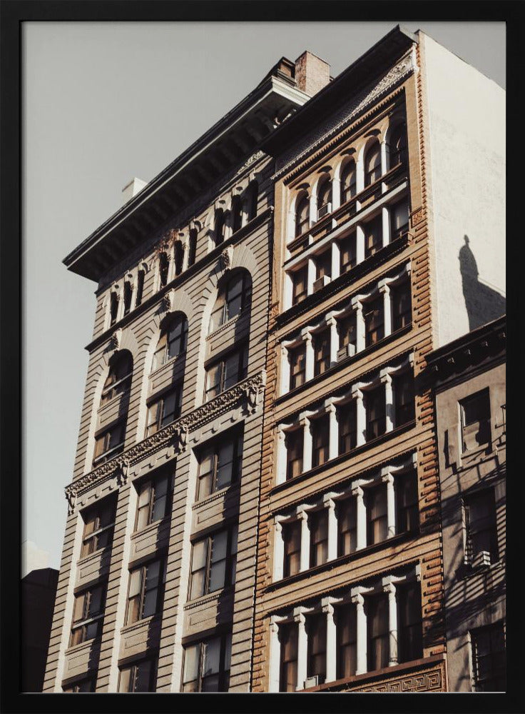 A low-angle, sunlit photograph of two ornate, multi-story historic buildings against a pale sky, framed in black. The detailed facades feature arched windows and decorative columns, with warm light creating strong shadows. Poster