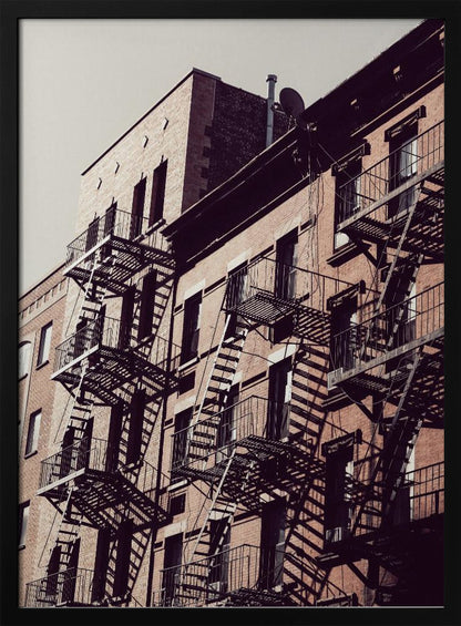 A low-angle photograph of a classic brick apartment building, with a complex pattern of black metal fire escapes casting long, dramatic shadows across the sunlit facade against a pale sky. Decor