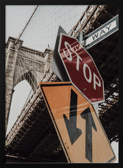A low-angle photograph of the Brooklyn Bridge with a collection of street signs in the foreground. A red stop sign and a yellow sign with two arrows are prominent against the architectural details of the bridge. Print