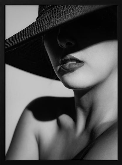 A dramatic black and white close-up photograph of a woman wearing a large, dark, wide-brimmed hat. The hat casts a deep shadow over the upper half of her face, obscuring her eyes and creating a sense of mystery. Only her nose, full lips, chin, neck, and shoulder are visible, highlighted by a strong light source. Decor