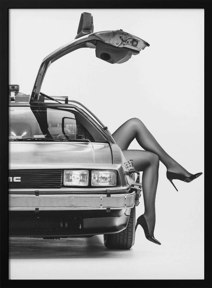 A black and white studio photograph of a DeLorean car with its gull-wing door open, featuring a woman's legs in black stockings and stilettos elegantly emerging from the passenger side. Decor