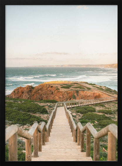 A wooden staircase and walkway descends a green, scrub-covered hill towards a rocky point overlooking a churning blue ocean under a pale, hazy sky. Print