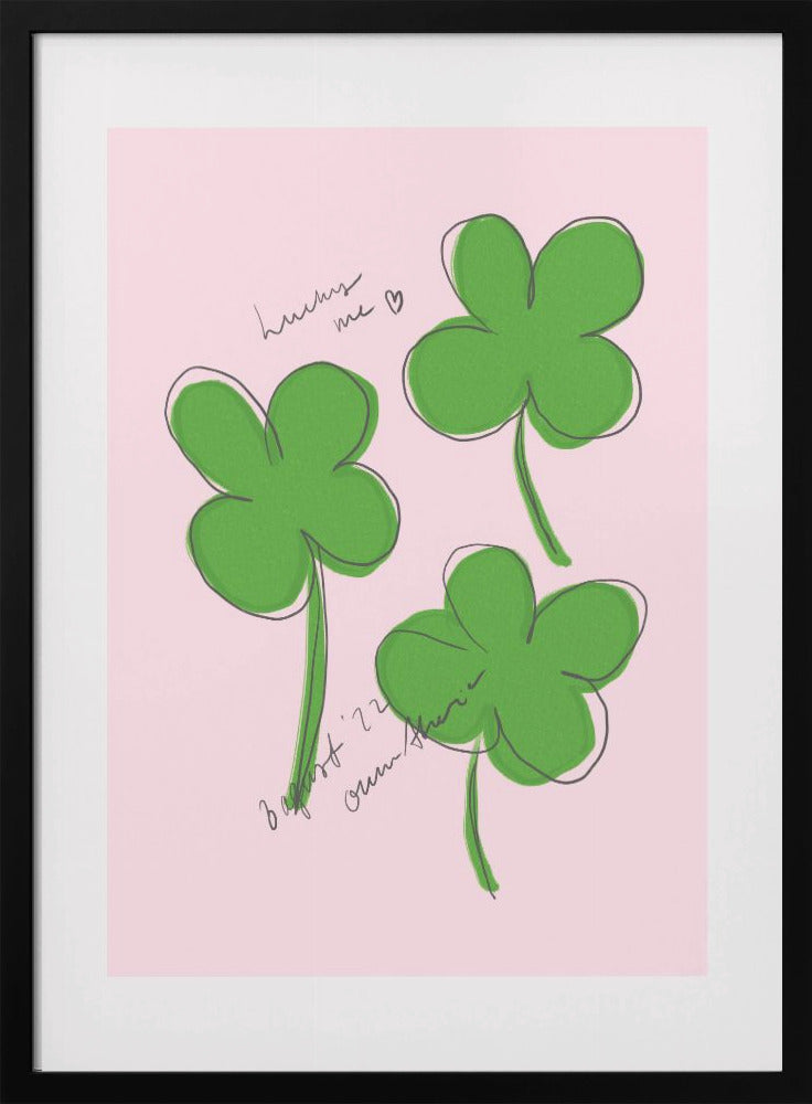 A framed art print featuring three hand-drawn four-leaf clovers in green with a loose black outline, set against a solid light pink background. Cursive handwriting on the print reads 'lucky me ♡' and includes a signature. Artwork